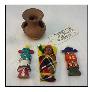 Puede incluir: Un conjunto de tres muñecas nativas americanas, una envuelta en una manta de cuadros rojos y negros, otra con un atuendo azul y naranja, y otra con un atuendo verde y blanco. También se incluye una pequeña maceta de terracota con cuatro agujeros. Las muñecas están hechas a mano y tienen un aspecto rústico y tradicional. La etiqueta dice: "To say I favorite time to write is around like a few excuse, but that's the truth, and to help with that I filled this paper with sill my this paper"
