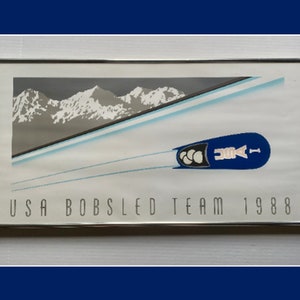 Puede incluir: Un póster enmarcado con una ilustración azul y blanca de un trineo de bobsleigh bajando por una pista con montañas en el fondo. El texto "USA Bobsled Team 1988" está impreso debajo de la ilustración.
