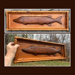 May include: Wooden wall art featuring a carved fish in a rectangular frame. The fish is a dark brown color, and the frame is a lighter brown with a rope-like border. The artwork is displayed outdoors.