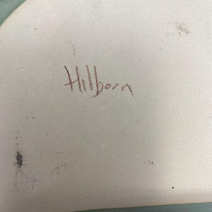 May include: A white ceramic plate with a blue and green glaze. The plate is signed "Hillbern" in brown ink.