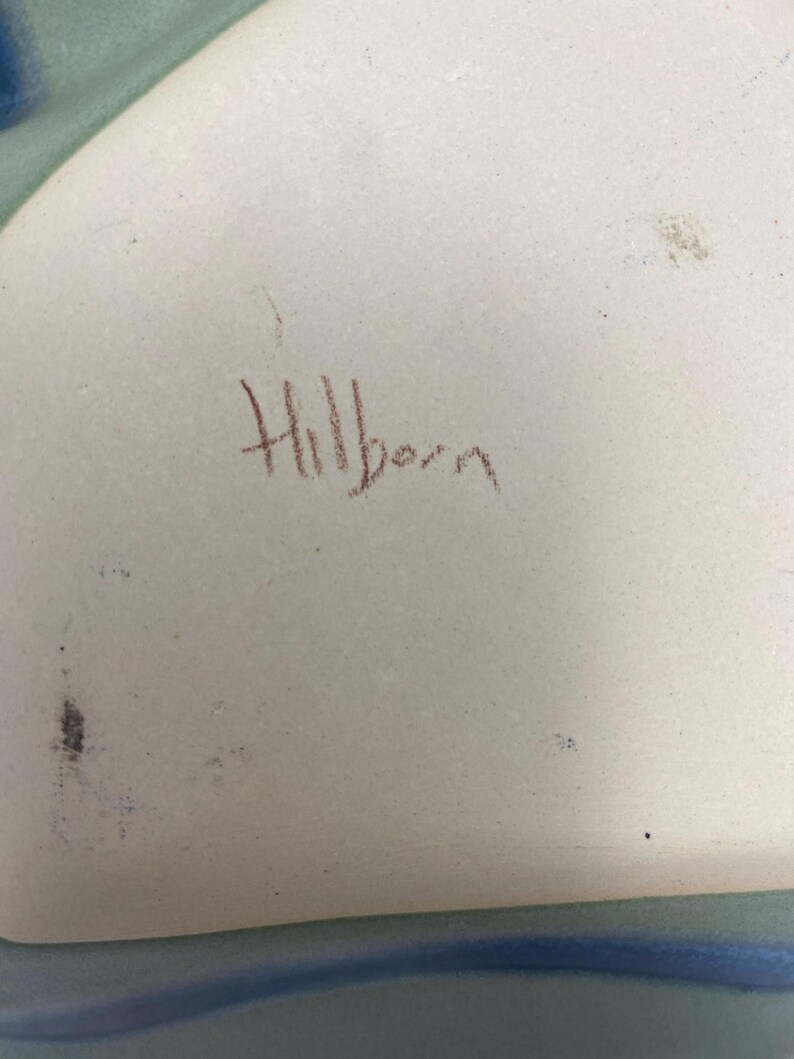 May include: A white ceramic plate with a blue and green glaze. The plate is signed "Hillbern" in brown ink.