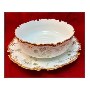 Puede incluir: Un cuenco de porcelana blanca con borde festoneado, sobre un platillo a juego. El juego presenta ribetes dorados y delicados motivos florales en tonos verdes y marrones. El fondo es rojo liso.