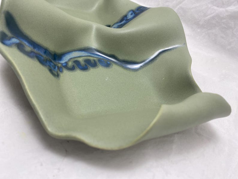 May include: A green ceramic dish with a wavy edge and blue and white glaze. The dish has a unique, organic shape.