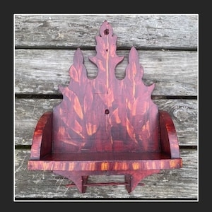 May include: A handcrafted, dark red wooden shelf with a decorative, flame-like top. The shelf has a small, curved front and a lower shelf with a small rod. The wood grain is visible, adding to its rustic charm.