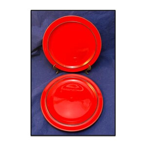 Op de afbeelding: Twee rode keramische borden met gouden rand. Eén bord staat op een goudkleurige standaard. De borden hebben een glanzende afwerking en staan tegen een donkerblauwe achtergrond.