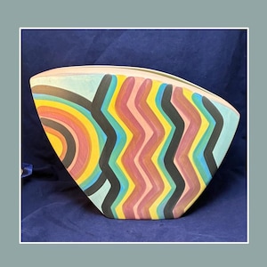 Puede incluir: Un jarrón de cerámica abstracto y colorido con una forma única. El jarrón presenta un diseño de arcoíris en un lado y un patrón en zigzag en rosa, negro y amarillo en el otro. El jarrón mide aproximadamente 30 cm de ancho y 20 cm de alto.