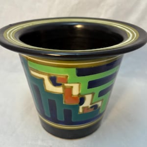 Puede incluir: Una maceta de cerámica con un borde ancho y acampanado de color base negro. El exterior está decorado con un patrón geométrico en tonos verde brillante, azul oscuro, crema y naranja quemado, con anillos concéntricos en el borde superior.
