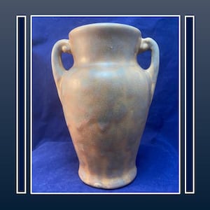 May include: A vintage, two-handled vase with a wide body and a narrow neck. The vase has a muted, earthy color palette with shades of brown and green. The handles curve gracefully from the neck to the body. The vase is likely made of ceramic or pottery.