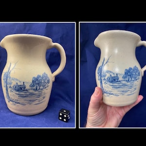 以下が含まれることがあります： 側面に田舎の風景が描かれた青と白の陶器のピッチャー。ピッチャーは口が広く、取っ手があります。