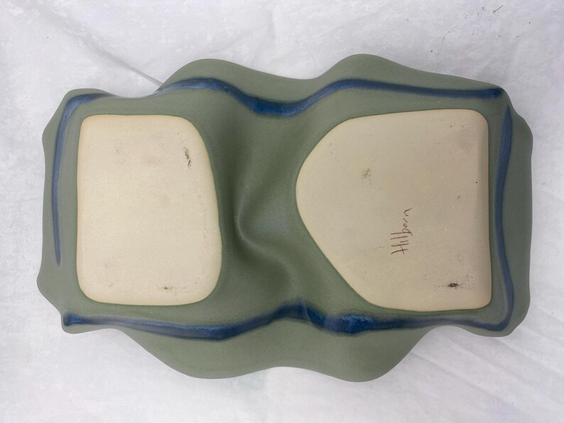 May include: A green ceramic serving dish with two indented sections. The dish has a blue glaze around the edges and the word "Hilltop" is written in brown ink on the bottom.