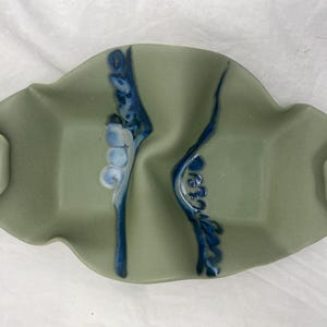 May include: A green ceramic serving dish with two divided sections. The dish has a wavy edge and is decorated with blue and white floral designs.