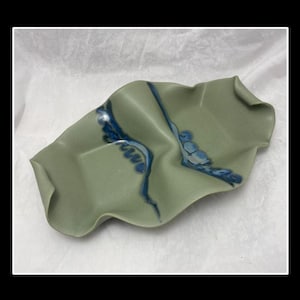 Puede incluir: Un plato de servir de cerámica verde con una forma ondulada y orgánica. El plato tiene un diseño de esmalte azul y blanco.