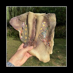 May include: An abstract ceramic sculpture with a textured, organic form. The piece features a blend of beige, brown, and gold and pink accents. The sculpture is held in a hand, showcasing its intricate details and artistic design. A unique art piece.