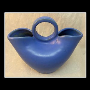 May include: A blue ceramic vase with a unique design. It features a central handle and two side openings. The vase has a smooth, matte finish and a rounded shape, suitable for floral arrangements or decorative display.