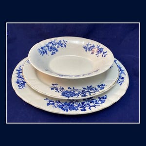May include: A stack of three white ceramic dishes with blue floral patterns. The set includes a shallow bowl and two plates, all with a gold trim. The dishes are arranged on a dark blue surface.