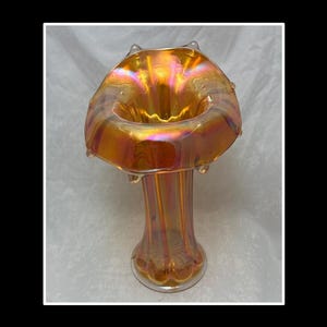 May include: An iridescent orange art glass vase with a flared, ruffled rim and a tall, slender stem. The vase has a vibrant, shimmering finish, showcasing hues of gold, orange, and purple. The vase is approximately 10 inches tall.