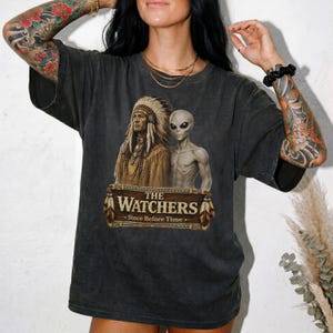 May include: A charcoal grey short-sleeve t-shirt featuring a graphic print of a person in traditional indigenous attire standing next to a grey alien. Below the figures is a banner with the text THE WATCHERS and the phrase Since Before Time.