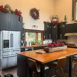 Può includere: Una cucina moderna con armadietti neri e un frigorifero in acciaio inox. La cucina è decorata per Natale con stelle di Natale rosse e bianche, ghirlande e festoni. L'isola della cucina ha un piano di lavoro in legno e sgabelli da bar.
