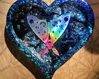 May include: Heart-shaped trinket dish with a holographic butterfly design. The dish has a dark blue and black interior with a shimmering, iridescent border. The butterfly features rainbow colors and celestial details.
