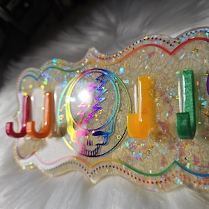 May include: A decorative wall-mounted key holder with a colorful design. The holder features the word "JJJ" in red, orange, and yellow, along with a Grateful Dead skull and lightning bolt graphic. The background is a glittery, iridescent resin.