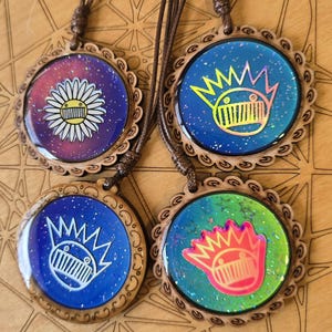Könnte beinhalten: Vier Holzketten mit runden Anhängern in verschiedenen bunten Designs. Die Anhänger haben eine glitzernde Oberfläche und zeigen eine Comicfigur mit einer Krone und einem Mund mit Zähnen. Ein Anhänger zeigt eine Gänseblümchen.