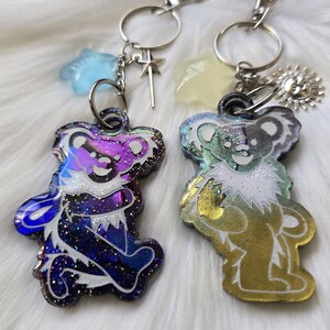 May include: Two iridescent glitter bear keychains with silver key rings. The bear on the left is a dark blue, purple, and pink color. The bear on the right is a green, gold, and yellow color.