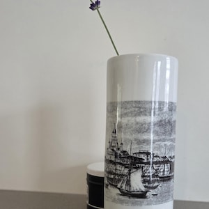 Peut inclure: Un vase cylindrique en céramique blanche orné d'une illustration imprimée en noir et blanc représentant un port avec des voiliers et des bâtiments. Un brin de lavande violette est placé dans le vase, dépassant de l'ouverture supérieure.