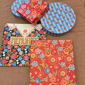 Peut inclure: Une collection de boîtes décoratives et un album carré avec des motifs floraux vibrants en rouge, bleu et vert. Les articles comprennent une boîte carrée rouge à fleurs, une boîte ronde bleue à rayures florales, une petite boîte ronde bleue à fleurs et un album carré avec le mot MEMORIES imprimé sur la couverture.