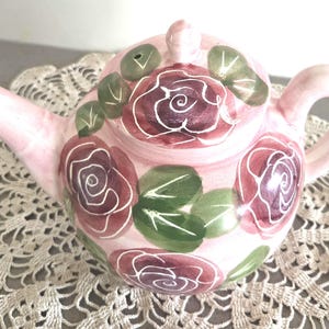 Peut inclure: Une théière en céramique rose clair ornée de roses rouge foncé et de feuilles vertes peintes à la main. La théière comprend un couvercle assorti avec un petit bouton, une anse incurvée et un bec verseur effilé. Le motif floral couvre le corps.