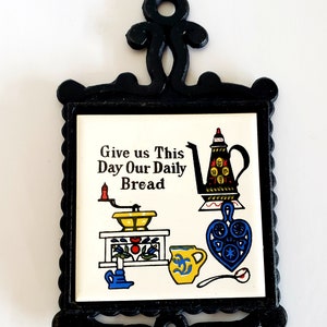 May include: Black cast iron pot holder with a white ceramic tile featuring a colourful illustration of a coffee grinder, teapot, and mug with the text "Give us This Day Our Daily Bread".