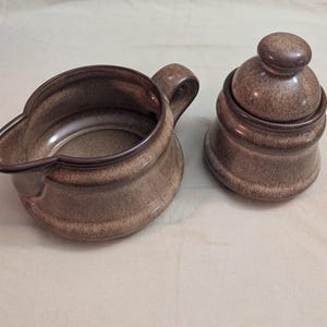 May include: A brown ceramic creamer with a handle and a matching sugar bowl with a lid. The set has a speckled finish and a dark brown rim. The creamer has a spout for easy pouring. The sugar bowl has a rounded shape.