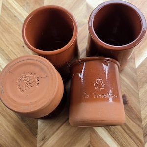 Puede incluir: Tres macetas de terracota con una sección superior lisa y esmaltada y una base mate sin esmaltar. Una maceta está invertida para mostrar el texto en relieve WWW.LAFERMIERE.COM. Otra maceta presenta el texto en relieve la fermière y un logo.