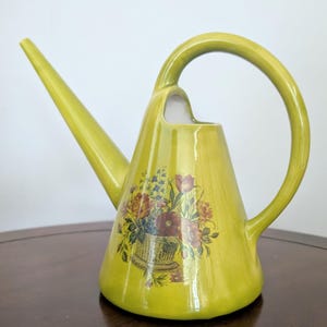 May include: A chartreuse ceramic watering can with a long, slender spout and a high, arched handle. The body features a central decal of a woven basket overflowing with various colourful flowers, including red, orange, and blue blossoms with green leaves.