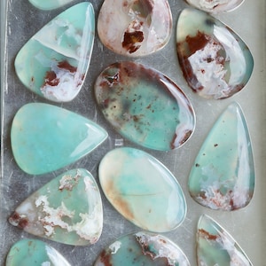 May include: A collection of polished gemstones in various shapes, including teardrop and oval. The stones feature a mix of pale green, white, and brown colors, with a translucent appearance. The arrangement is on a silver surface.