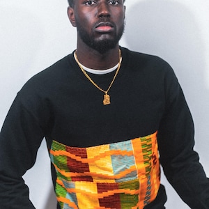 Könnte beinhalten: Ein schwarzes Langarmshirt mit einem bunten, gemusterten Einsatz über der Brust. Das Muster ist eine Mischung aus Orange, Gelb, Grün und Rot. Das Shirt hat eine goldene Kettenkette mit einem Anhänger.
