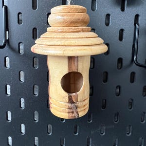 May include: A handcrafted wooden birdhouse with a layered roof design and a circular opening. The birdhouse is made of light-colored wood with darker wood accents and is suspended by a thin silver wire. The overall design is reminiscent of a pagoda.