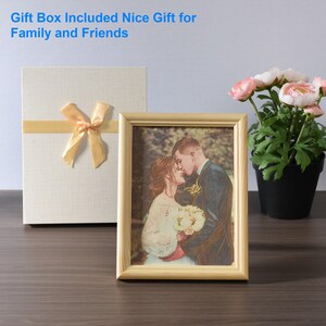 May include: A framed photo of a couple in a forest setting. The photo is in a light wood frame and is sitting on a wooden table. There is a gift box with a yellow ribbon and a bouquet of pink flowers in the background.