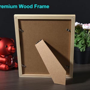 May include: A wooden picture frame with a light brown finish. The frame is standing on a brown surface with a red teddy bear in the foreground. The frame has a cardboard backing and a kickstand.