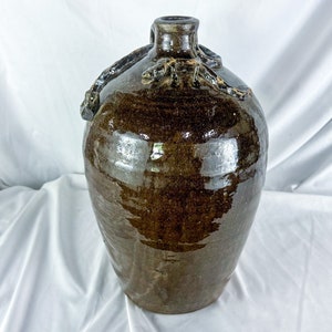 May include: A brown ceramic jug with a wide mouth and a handle. The jug has a dark brown glaze and a textured surface. The handle is shaped like a twisted vine.