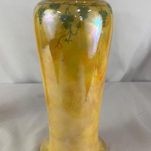 May include: A tall, iridescent yellow vase with a wide base and a narrow neck. The vase features a painted design of green grape vines and clusters around the top. The vase has a glossy finish and a pearlescent sheen.