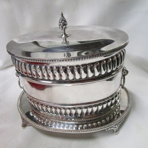 May include: A silver plated cake stand with a domed lid and ornate detailing. The stand has a round base with three feet and a decorative knob on the lid.