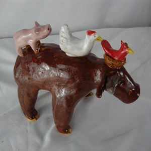 May include: A whimsical ceramic sculpture featuring a brown dog-like figure with a pink pig, a white chicken, and a red bird perched on its back. The sculpture has a glossy finish and is set against a white background.