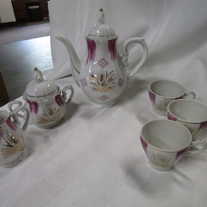May include: A white tea set with gold and pink accents. The set includes a teapot, creamer, sugar bowl, and four cups. The cups and teapot have a floral design with gold accents.