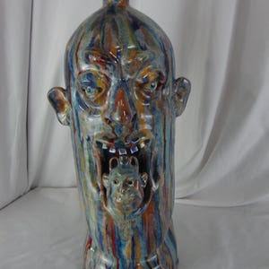 May include: A tall, ceramic vase with a face design. The vase features a grotesque face with blue eyes, an open mouth, and a smaller face inside the mouth. The vase is painted with vertical streaks of blue, orange, and brown.