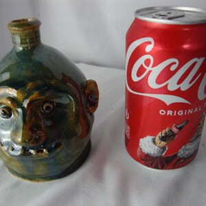 May include: A ceramic jug with a face design, featuring a smiling expression, is next to a red Coca-Cola can. The jug has a green and brown glaze. The Coca-Cola can has the word "Coca-Cola" in white script.