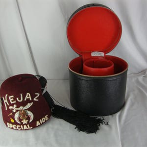 May include: A burgundy fez hat with gold and silver embroidery, the text "HEJAZ SPECIAL AIDE" is visible. A black, round hat box with a red interior is open next to the hat. The hat has a black tassel.