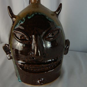 May include: A brown glazed ceramic face jug featuring a wide, toothy grin, pointed ears, and two horns protruding from the top. The vessel has a glossy finish with subtle blue-green highlights around the facial features and a central spout.