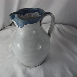 May include: A handmade ceramic pitcher with a white body and a blue rim and handle. The pitcher has a rounded shape and a spout for pouring. The handle is curved and easy to grip. This decorative pitcher is perfect for serving water or other beverages.