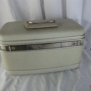 May include: A vintage, light-colored vanity case with a textured finish. The rectangular case has a silver-toned handle and trim, with a secure latch closure. The case is designed for travel and storage of cosmetics and personal items.