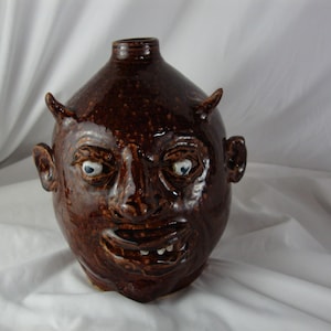 May include: A brown ceramic jug with a face sculpted on the front. The face has a wide mouth, a large nose, and two small horns on the top of the head. The eyes are painted blue.